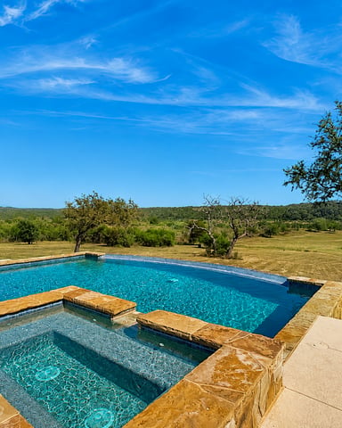 Swimming pool and hot tub with stone edges overlooking rural landscape with scattered trees and clear blue sky