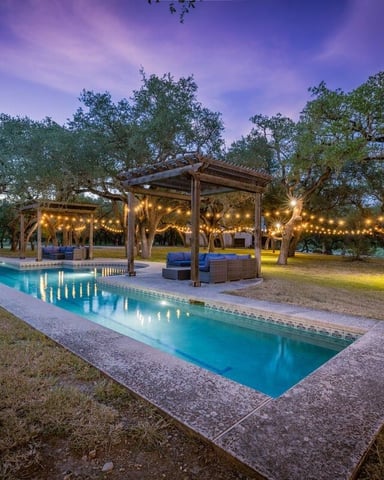 Rectangular swimming pool with concrete deck surrounded by mature oak trees, wooden pergola with string lights, and lounge seating at dusk.