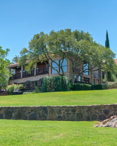 Stone and timber house with large windows set on manicured lawn, surrounded by mature trees and flowering shrubs