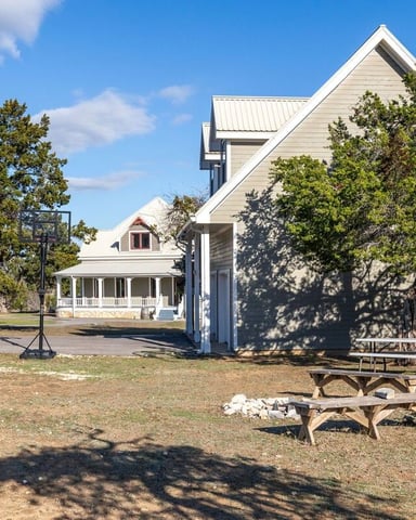 Swimming pool with stone deck facing two-story white house under large oak trees, grill and seating area visible