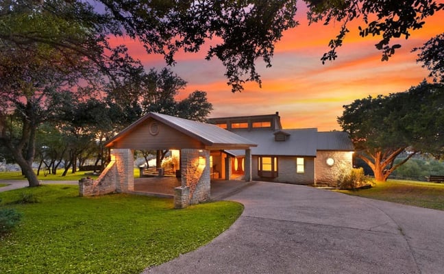 Modern stone house with covered porch at sunset, surrounded by large oak trees and manicured lawn